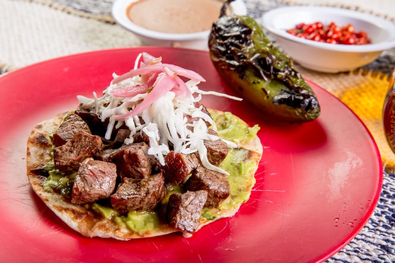 A red plate holds a tortilla topped with steak, guacamole, shredded cabbage, and pickled onions.