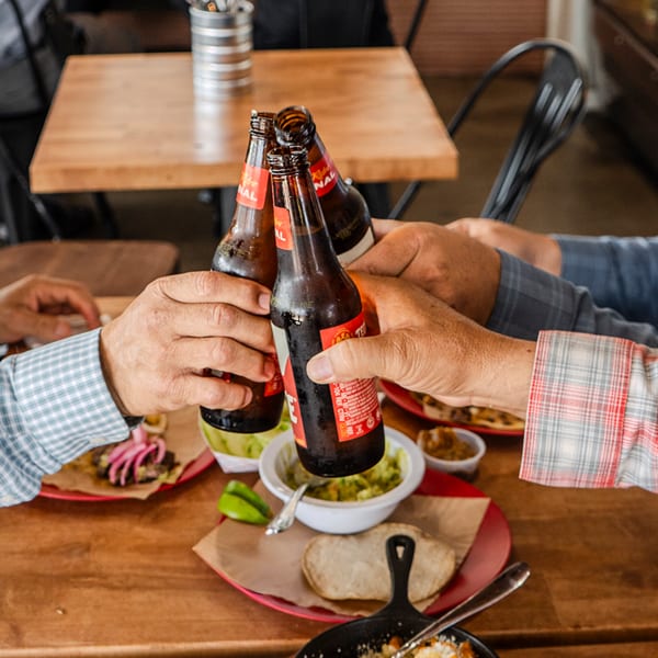 Four people clinking beer bottles together over a table filled with Mexican food and drinks.