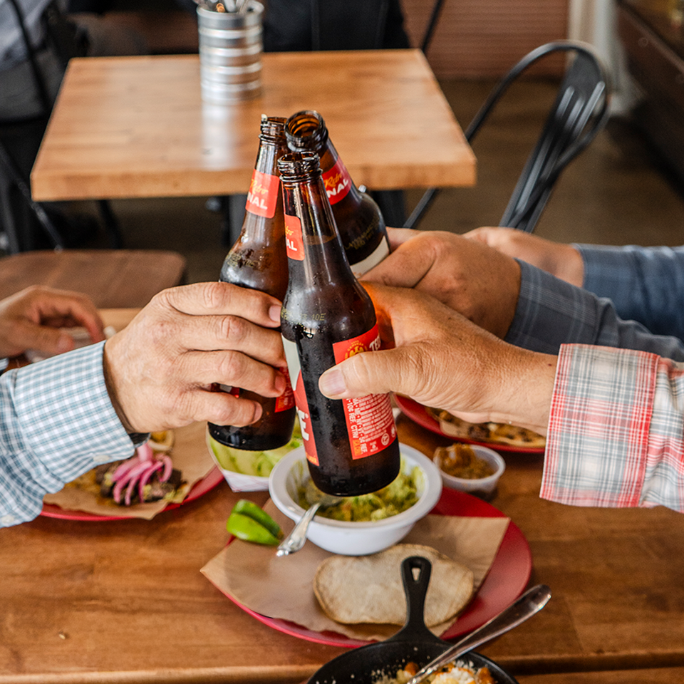 Four people clinking beer bottles together over a table filled with Mexican food and drinks.