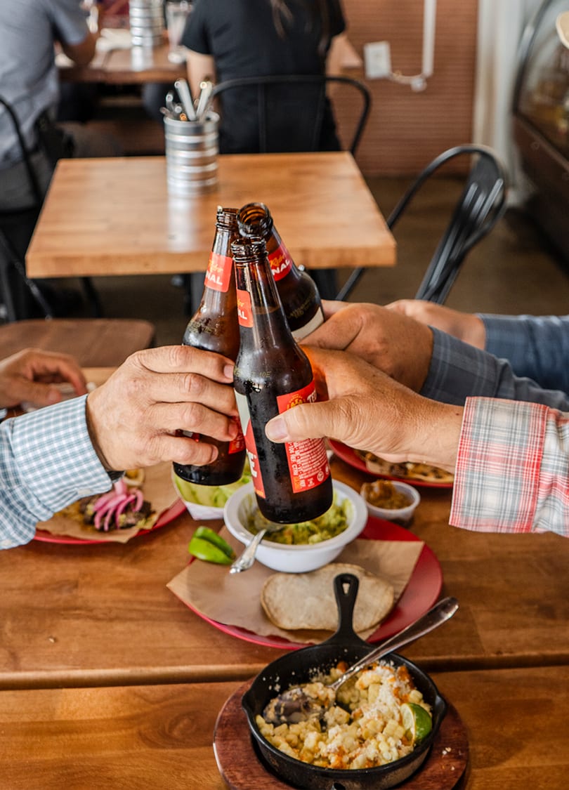 Four people clinking beer bottles together over a table filled with Mexican food and drinks.
