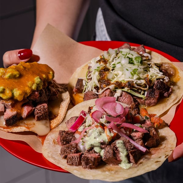 A person holds a red plate with three steak tacos, each with different toppings.