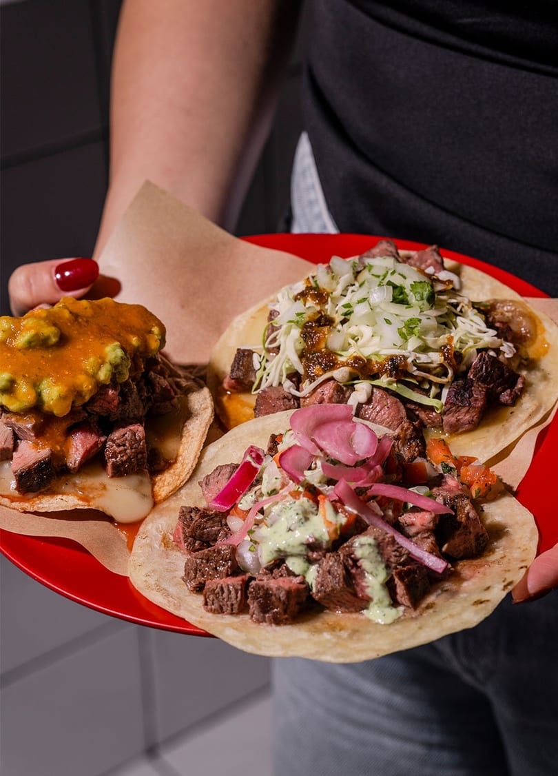 A person holds a red plate with three steak tacos, each with different toppings.