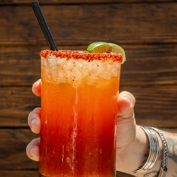 Hand holding a vibrant orange and red cocktail with a chili-lime rim, straw, and lime wedge.