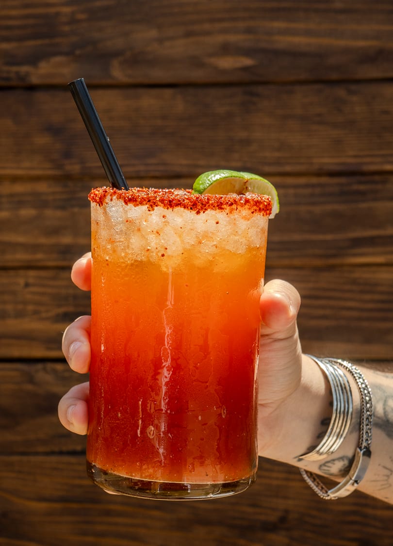Hand holding a vibrant orange and red cocktail with a chili-lime rim, straw, and lime wedge.