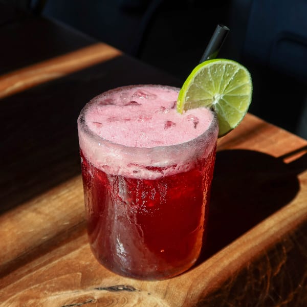 A vibrant red cocktail with a foamy top, garnished with a lime slice and black straw on a wooden table.