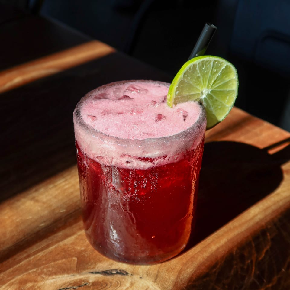 A vibrant red cocktail with a foamy top, garnished with a lime slice and black straw on a wooden table.