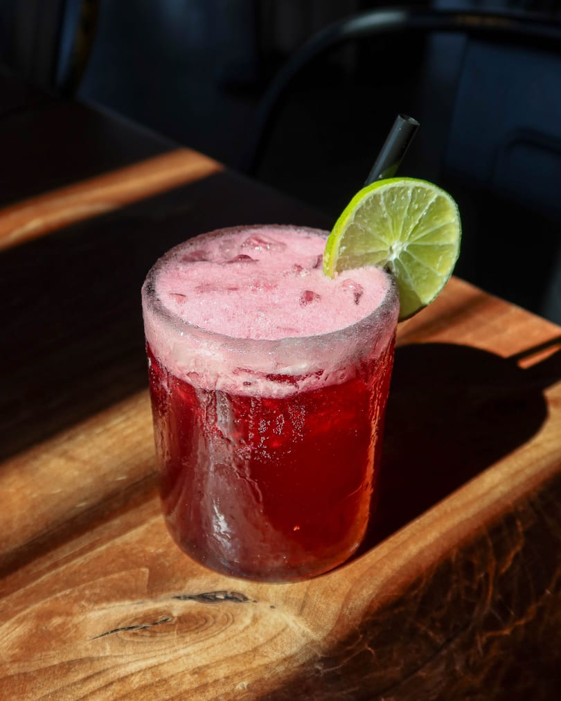 A vibrant red cocktail with a foamy top, garnished with a lime slice and black straw on a wooden table.