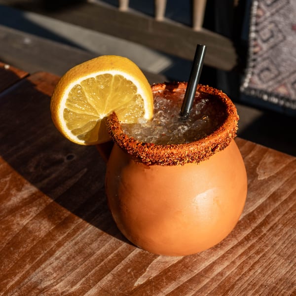 A clay mug with a chili-salted rim, filled with an iced drink and garnished with a lemon slice.