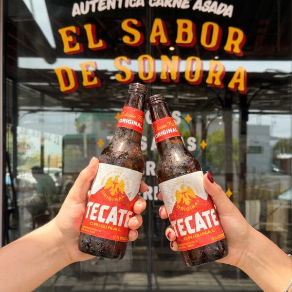 Two hands holding Tecate beer bottles, clinking them together in front of a restaurant window.