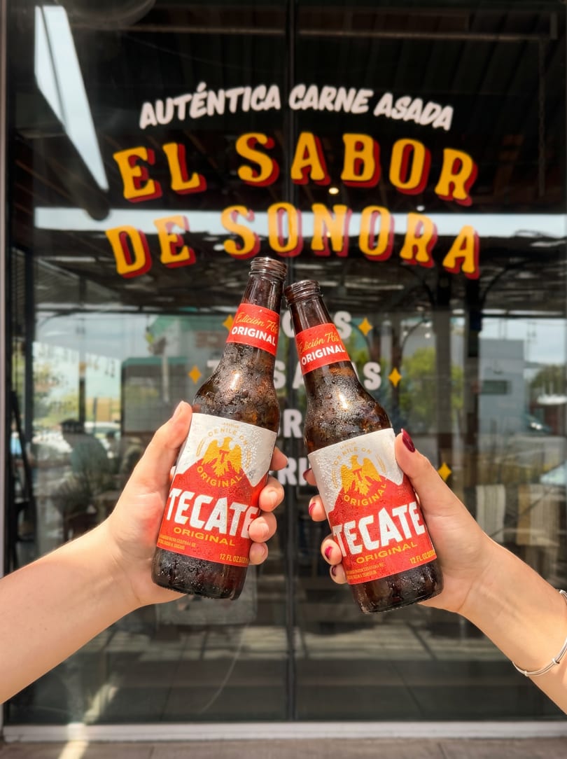 Two hands holding Tecate beer bottles, clinking them together in front of a restaurant window.