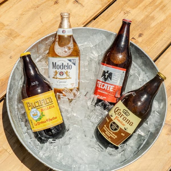 Four Mexican beer bottles, including Modelo, Pacifico, Tecate, and Corona, chilling in a galvanized bucket of ice.
