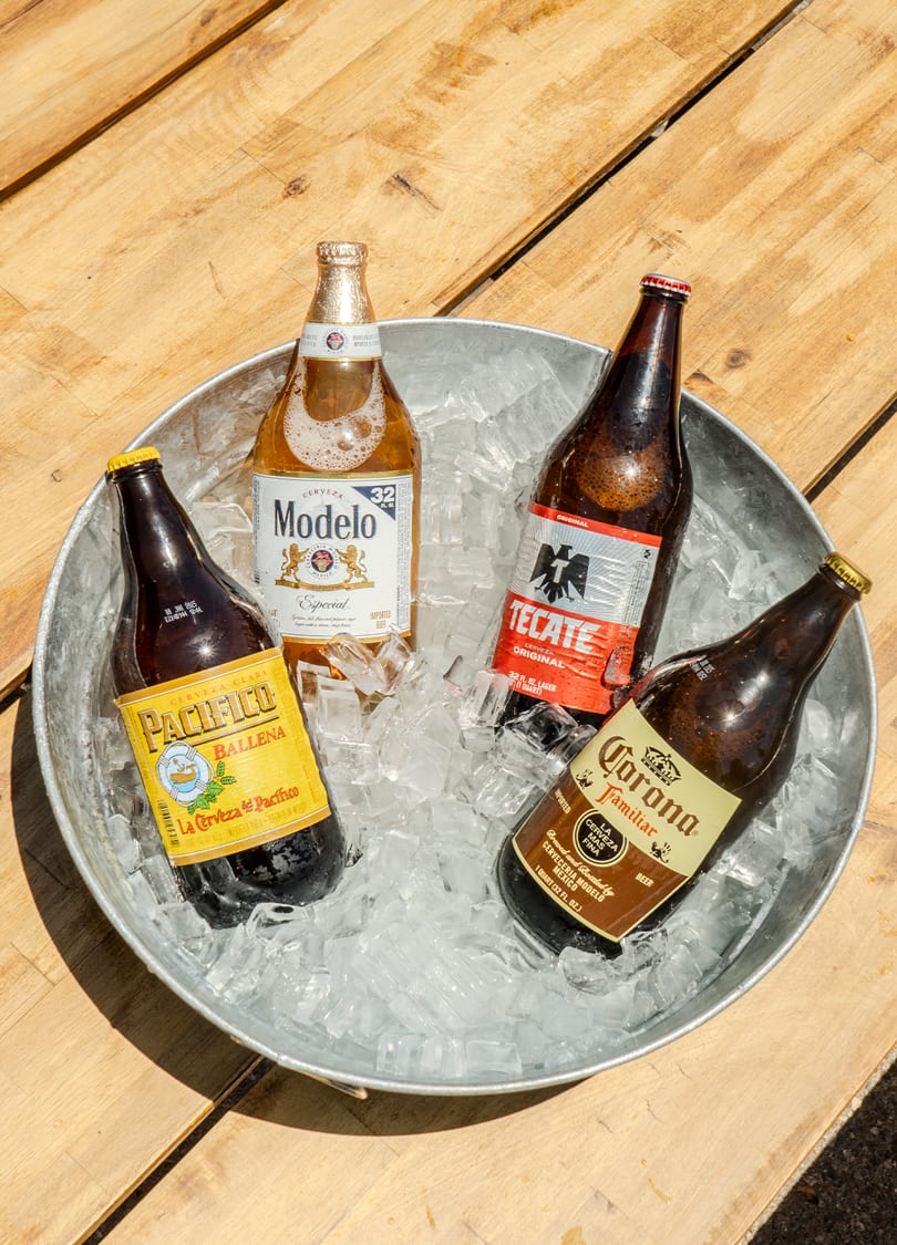 Four Mexican beer bottles, including Modelo, Pacifico, Tecate, and Corona, chilling in a galvanized bucket of ice.