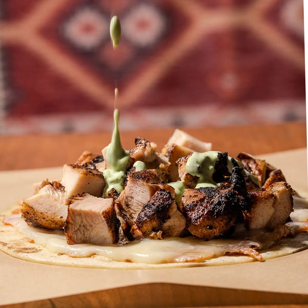 Green sauce drizzles onto a tortilla with grilled chicken and melted cheese on a wooden table.