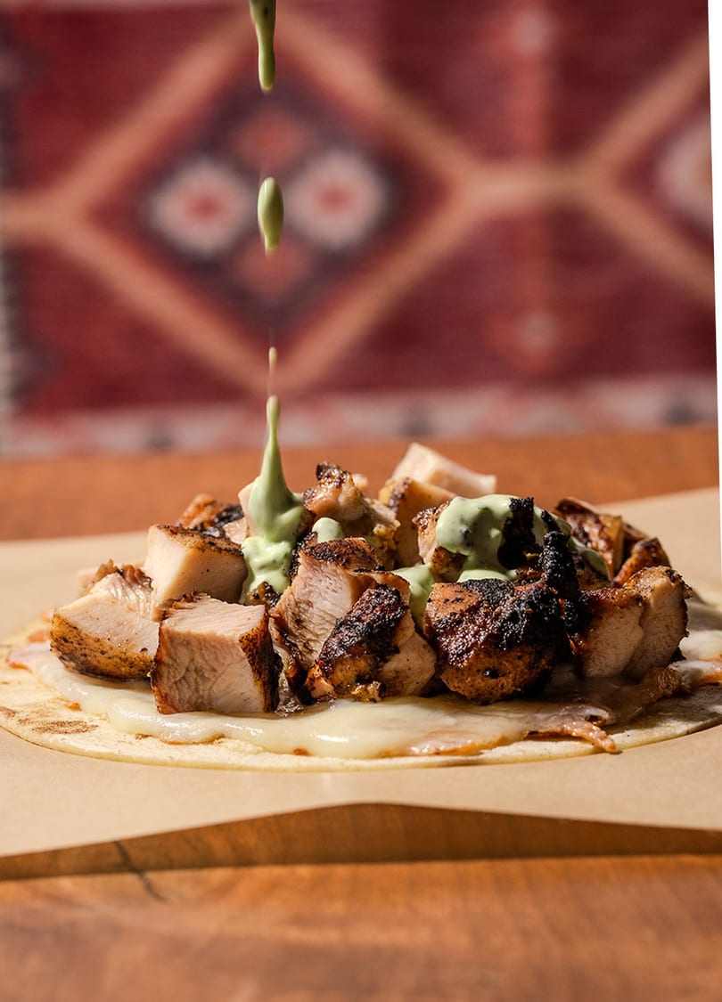 Green sauce drizzles onto a tortilla with grilled chicken and melted cheese on a wooden table.